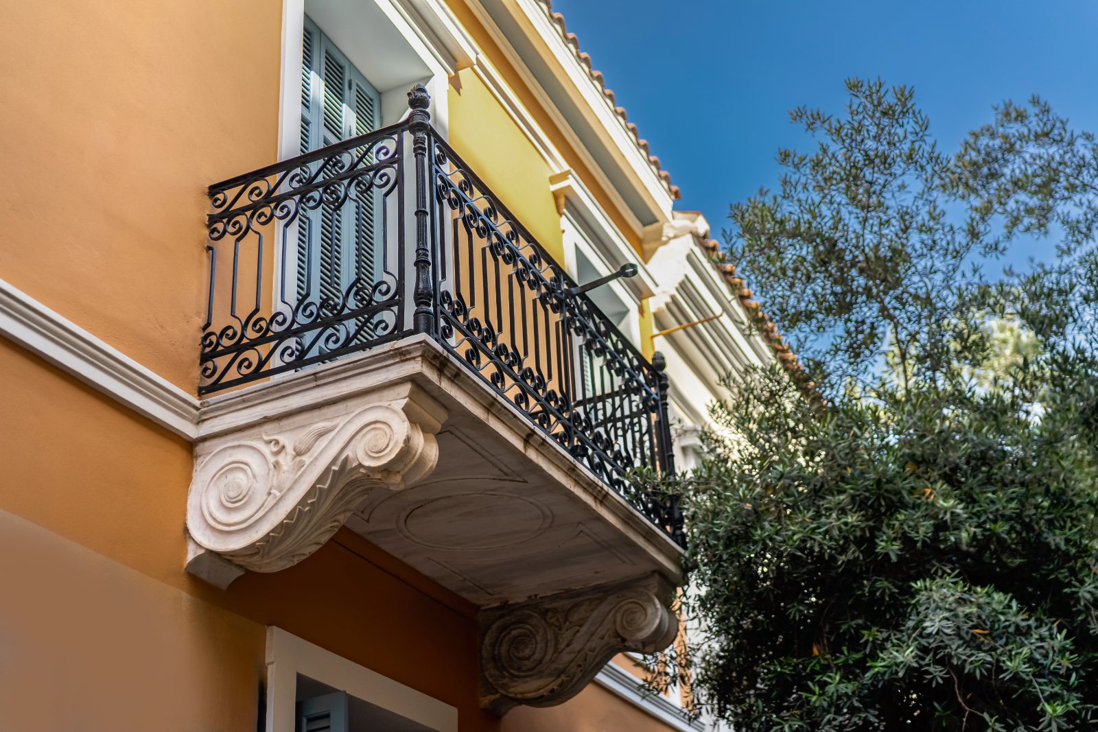 A neoclassical corner balcony in Exarchia, carved marble brackets and cast-iron railings against a blue sky