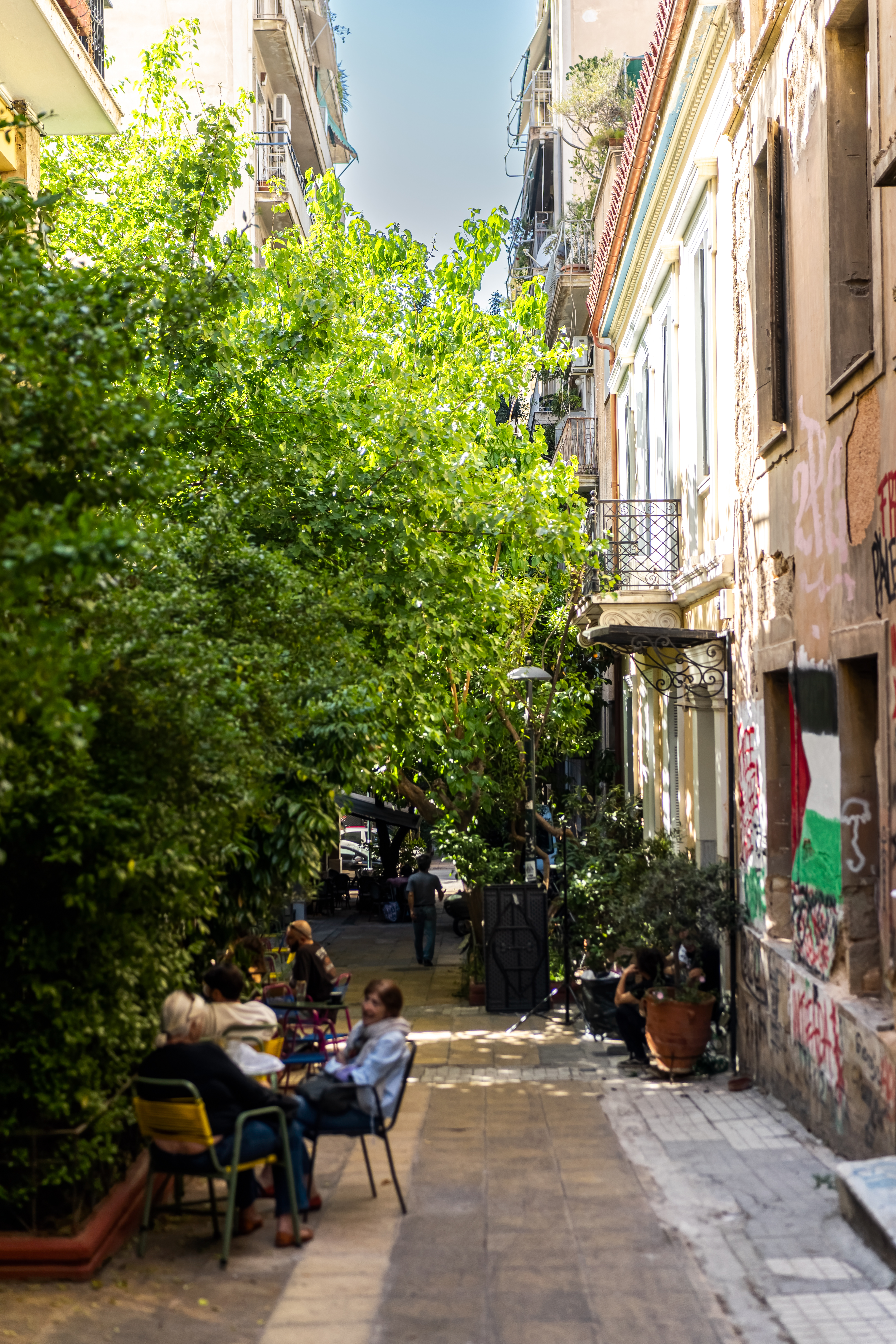 A shaded Exarchia side street, café chairs under the trees and graffiti on the wall opposite