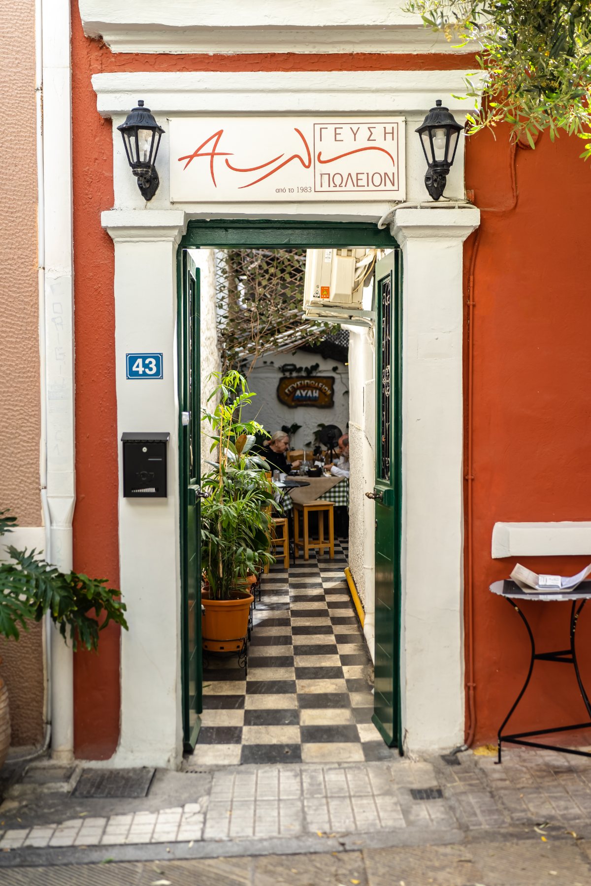 An open green door in a terracotta wall at number 43, a checkerboard-tiled passage leading through to a taverna courtyard